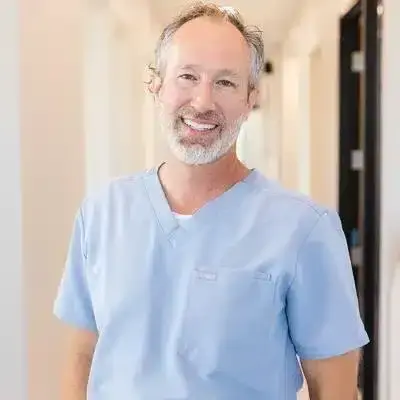 Male dentist in blue scrubs smiling in Queen Creek dental office hallway.