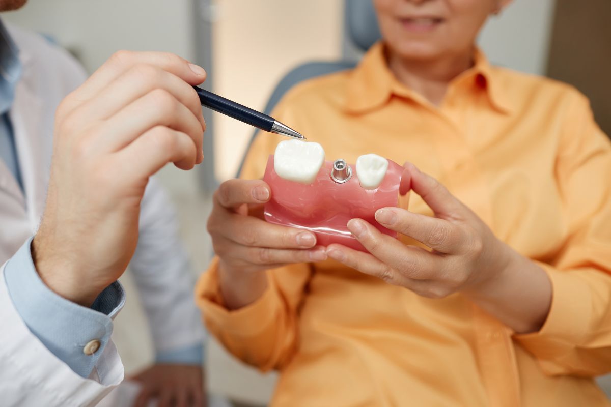 Dentist pointing to dental implant model while explaining recovery timeline to patient in yellow shirt.